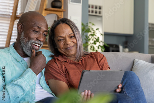 Senior couple sitting closely on light-gray sofa at home, looking at tablet together