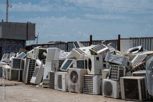 A massive pile of discarded and broken air conditioner compressor units sits outdoors in a junkyard under a blue sky.