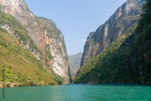 Majestic mountains frame turquoise river waters. Lush greenery blankets steep cliffsides. Clear blue sky above serene waterway. Natural landscape invites peaceful exploration
