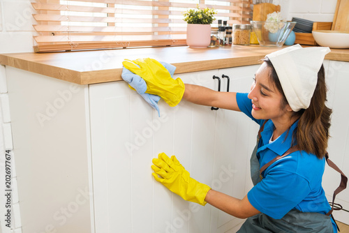 Smiling woman in rubber gloves cleaning a kitchen cabinet with a microfiber cloth, wearing an apron and headband in a bright home interior.