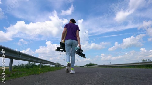 An adult woman in jeans walks along an asphalt road and holds roller skates. Her back is to the camera.