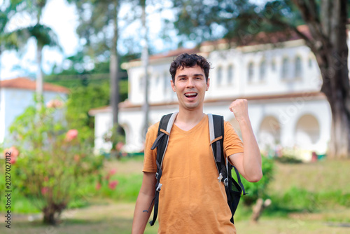 Excited Brazilian university student celebrating success on campus. He wears a backpack and raises his fist in victory, with a historic academic building and green trees in the background.
