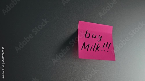 A sticky note with a shopping reminder is attached to a wall in an indoor location. The note emphasizes the importance of buying milk, showcasing a common task that many people need to remember