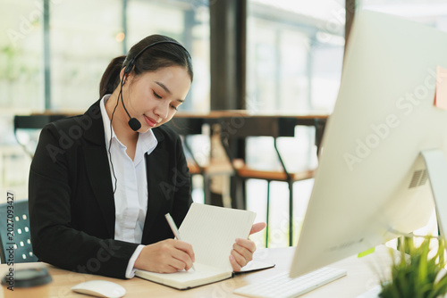Tableau sur toile Asian woman wearing a headset and formal attire, taking notes in a notebook whil