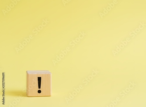 Single wooden cube displaying a black exclamation mark on a soft pastel yellow studio surface