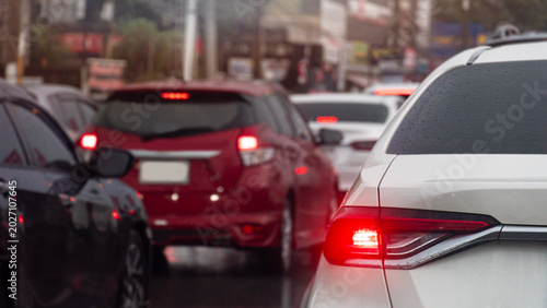 Vehicles are tightly packed in bumper to bumper traffic on a city street. A white car with its red brake light glowing is prominent on the right side of the busy urban frame. All wet from rain.