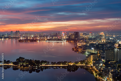 Evening aerial cityscape of West Lake and Thanh Nien Road featuring illuminated Hanoi skyline, reflective waters, and vibrant sunset colors.