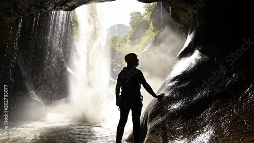 Man in silhouette stands by a roaring waterfall in a cave, ready for canyoneering adventure or rappelling darecation in nature.