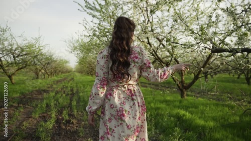 Young brunette in long floral dress with beautiful hairstyle walks through blooming garden and touches tree branch with her hand on spring day