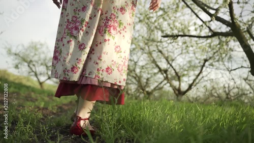 Young woman in red shoes and red and white floral dress walks through green grass among trees in spring. Close-up of legs.