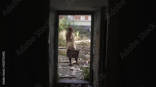 Young woman in beige coat, brown hat, boots and with suitcase stands in the doorway of ruined old building