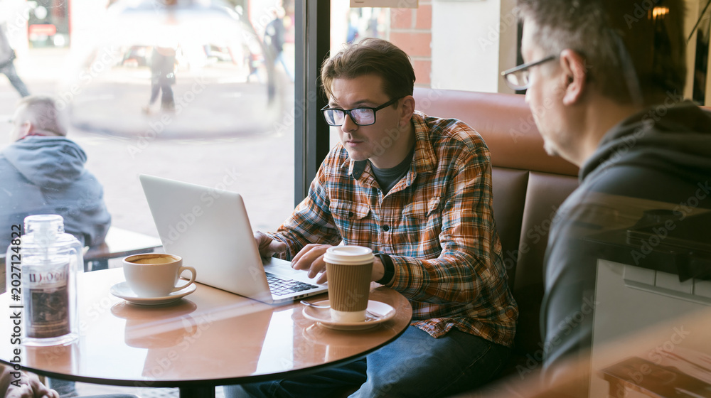 Fototapeta premium Nervous beginner entrepreneur opens a laptop for a first business meeting in a modest cafe while a client listens over coffee cups