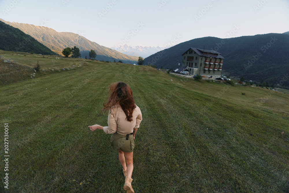 Fototapeta premium Woman walking across grassy hillside toward distant mountains, casual outdoor lifestyle, soft light, fresh air, expansive landscape, relaxed posture, natural colors.