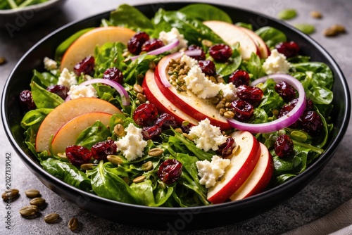 Close-up image of fresh green salad with apples, tangy goat cheese, sweet cranberries, thin red onion slices, and pepitas presented in a black bowl on a textured surface
