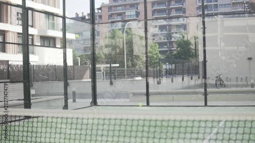 Empty padel court with balls flying toward the glass wall during a game or training session. Urban outdoor sports scene showing motion, impact and racket sport atmosphere without people