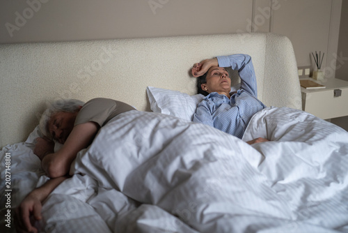 Vászonkép Frustrated senior woman struggling with night sleep because of husband snoring annoyingly at night, irritated female laying in bed with apathy and fatigue while man resting in deep sleep and snores