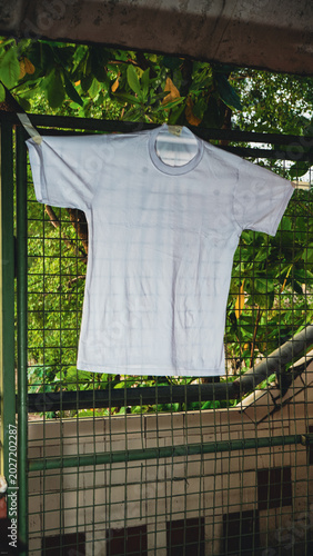 White t-shirt hangs on a metal fence. Green leaves frame the top of the shot. Background shows distant buildings and trees. Soft daylight casts gentle shadows. Image suggests a quiet, everyday moment