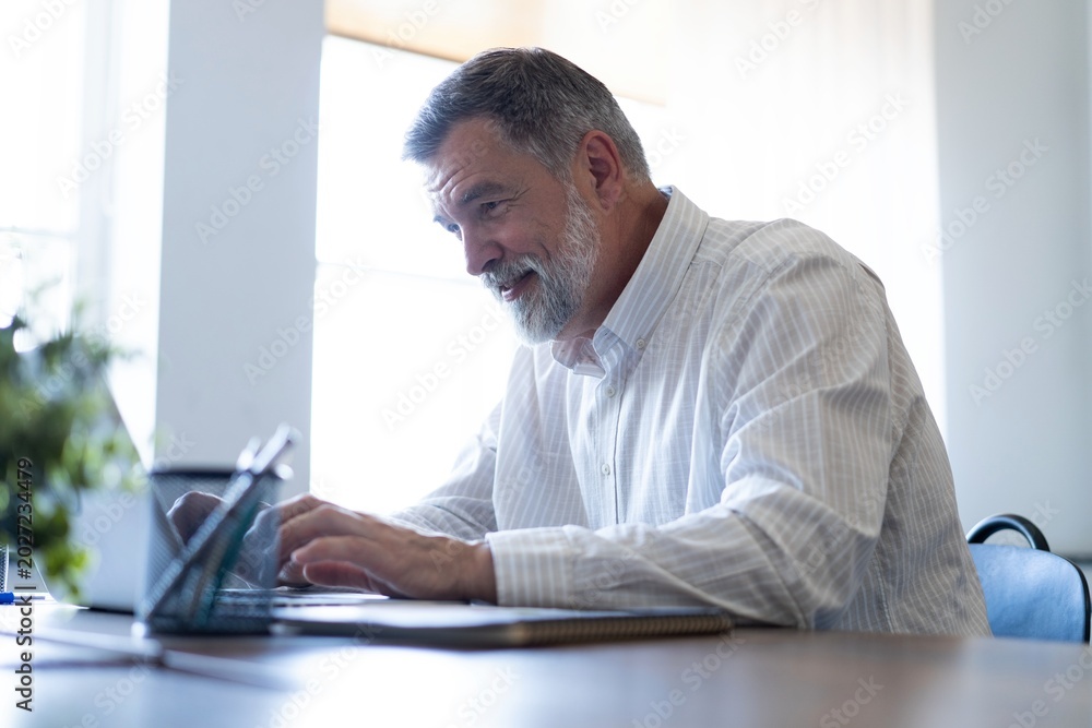 Fototapeta premium Smiling mature business man executive wearing shirt sitting at desk using laptop. Happy busy professional middle aged businessman investor working on computer looking away in office
