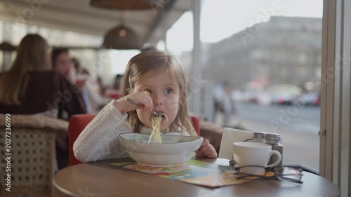 A beautiful, fair-haired little girl eats pasta in a restaurant. A child eats food with gusto on a street cafe terrace. Kid child holding fork in hand.