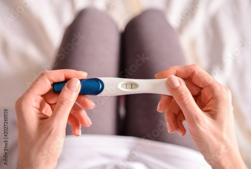 Adult female hands holding positive pregnancy test with two red lines, seated on bed, symbolizing conception, fertility, anticipation, and life change