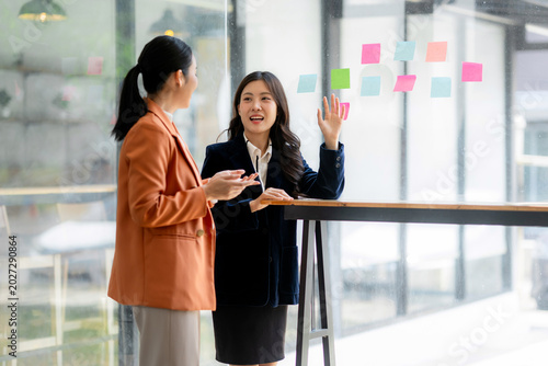 Two Asian businesswomen collaborating, discussing project plans, and organizing sticky notes on a glass board in a modern office