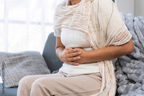 Woman sitting on sofa suffering from severe stomach ache and abdominal pain, holding abdomen due to digestive problem or period cramps