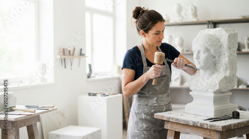 Female sculptor chiseling a classical bust in an art studio. Young woman carving a white stone statue with a mallet and chisel. Creative artistic process