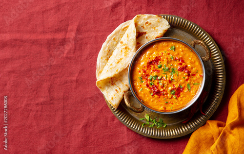 Rustic Indian cuisine: hearty dal dish with fluffy chapati, herb garnish, traditional serving style, warm textile backdrop. Deep red background. Copy space, close up, top view.