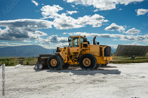 Heavy yellow front end wheel loader parked on a dusty white marble quarry surface under a gorgeous sunny blue sky dotted with fluffy clouds with solar panels and distant mountain range in view.