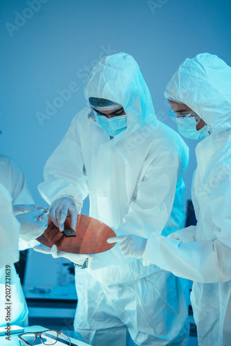 Engineers in cleanroom suits examine a silicon semiconductor wafer and microchip. Laboratory team conduct high tech research, microelectronics development, and hardware hardware manufacturing