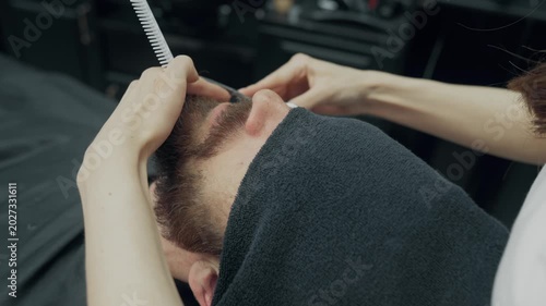 Professional barber trimming beard of male client with scissors and comb during grooming procedure in modern barbershop salon