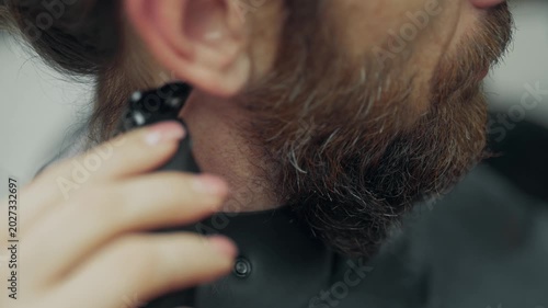 Close up of barber trimming neck hair with electric clipper during beard grooming procedure in modern barbershop