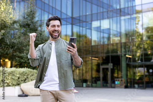 Photos Happy man looking at his smartphone, raising a fist in triumph, receiving excell