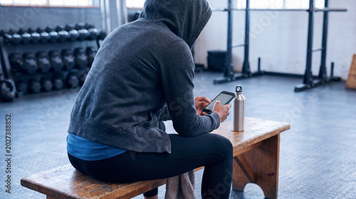 Konstfotografi Anonymous person in a hoodie sitting on a gym bench, engrossed in their smartphone with a water bottle nearby