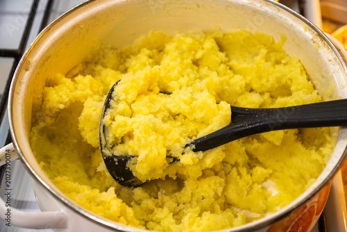 Slika na platnu High angle view of hot freshly boiled yellow potatoes mashed inside traditional enameled kitchen pot using black plastic hand masher tool during home cooking process