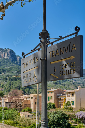 Wrought iron information sign for the historic tramway in Mallorca, showing directions with accessibility and stairs icons under a bright blue summer sky.