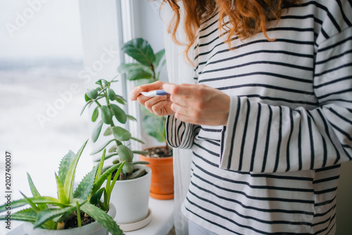 Woman standing next to a window with house plants, holding a pregnancy test while contemplating the potential life change and future family planning, symbolizing a personal health decision