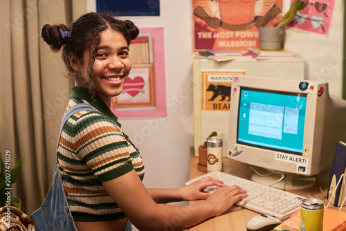Portrait of teenage girl using desktop computer at home desk, smiling at camera while typing, representing digital learning and youth technology. Useful for education or teen tech campaigns