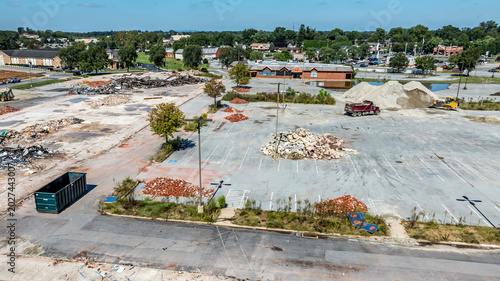 Workers are clearing a site with piles of rubble and dirt. The area is empty with scattered debris and a few vehicles. Nearby buildings are visible under a clear sky.