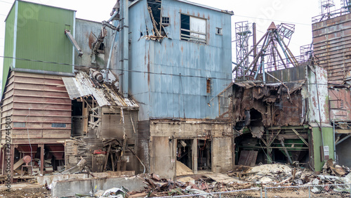 A large structure stands in an urban area showing signs of damage and decay. Parts of the building are crumbling and materials are strewn about the ground. The sky is overcast.