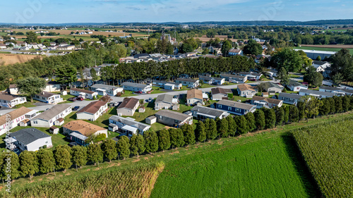 A collection of mobile, manufactured homes aligned in rows in a rural community. The houses are arranged in a neat pattern with trees and open fields nearby. Farms are visible in the distance.