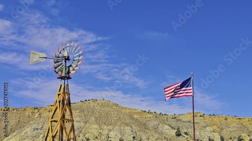 Old fashioned wind mill turning in the wind with American flag waving along side against blue sky with light clouds and buff collared hills and cliffs