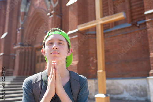 Teenage boy tourist praying with folded hands near catholic church cross