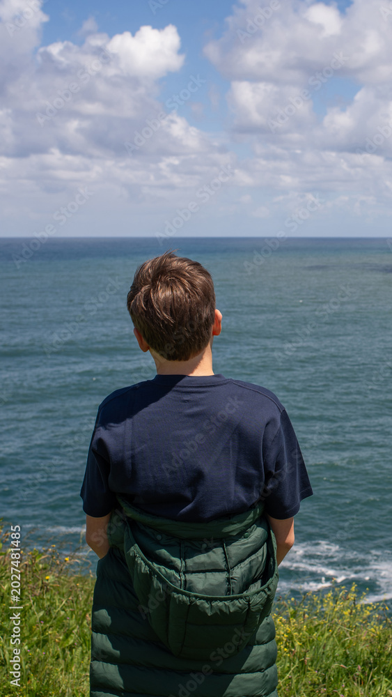 Fototapeta premium Teenage boy standing on coastal cliff and looking at ocean horizon in Asturias, Spain. Peaceful travel and nature scene with Atlantic sea view and dramatic cloudy sky.