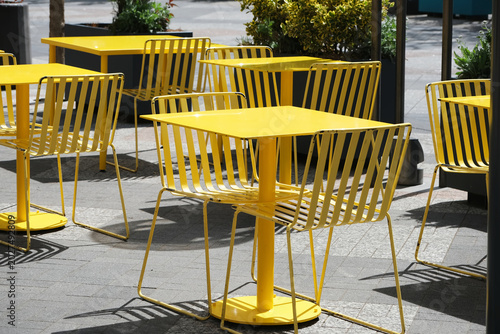 Urban cafe patio with bright yellow metal tables and wire chairs arranged on grey pavement. Modern bistro seating casting geometric shadows, empty outdoor dining and design scene.