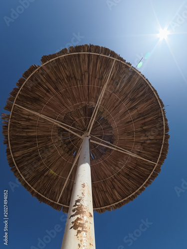 Low angle look up view of a straw beach umbrella against a clear blue sky with bright summer sun flare