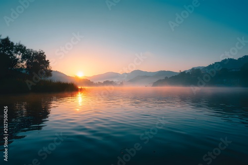 Ethereal sunrise over a tranquil lake with floating mist and silhouetted mountains in a hazy atmosphere