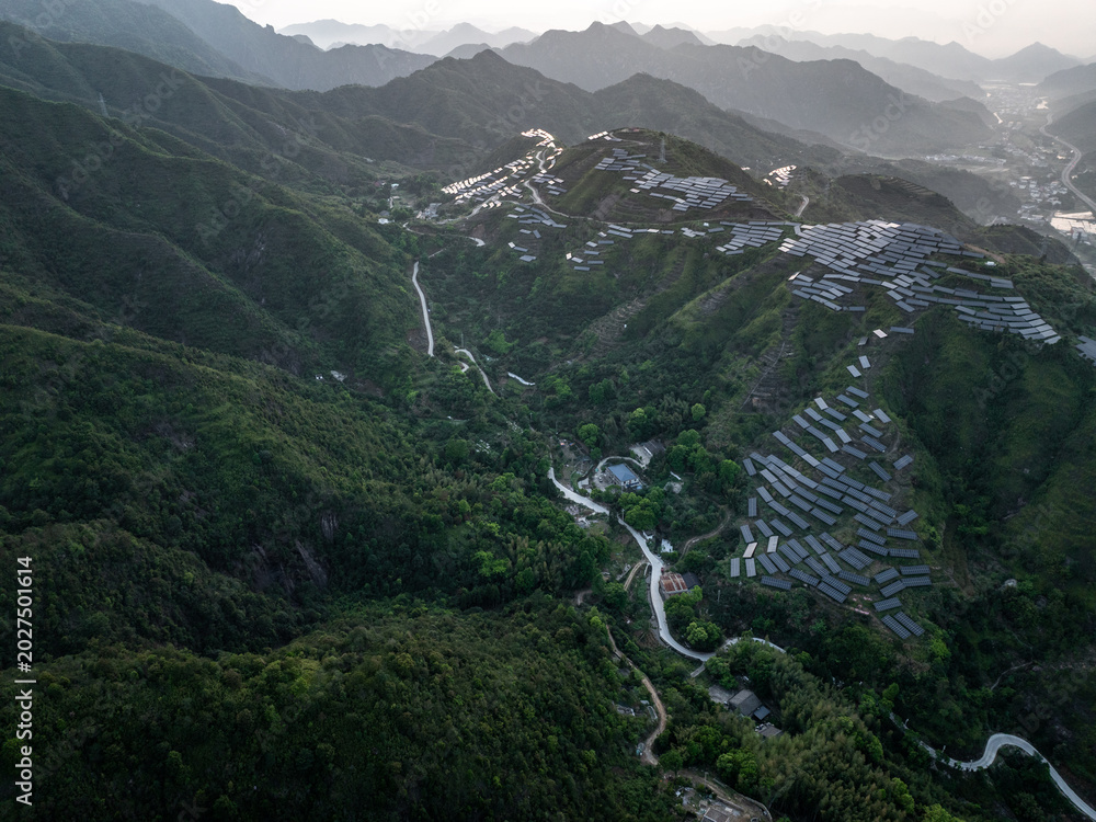 Fototapeta premium Aerial view of photovoltaic panels on terraced hills with winding roads and green forest landscape