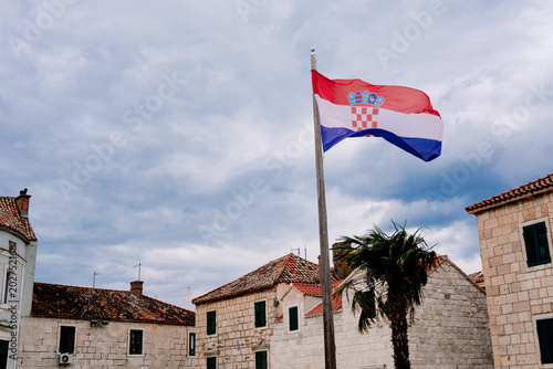 Croatian flag waves by ancient stone architecture & palm tree under an overcast sky