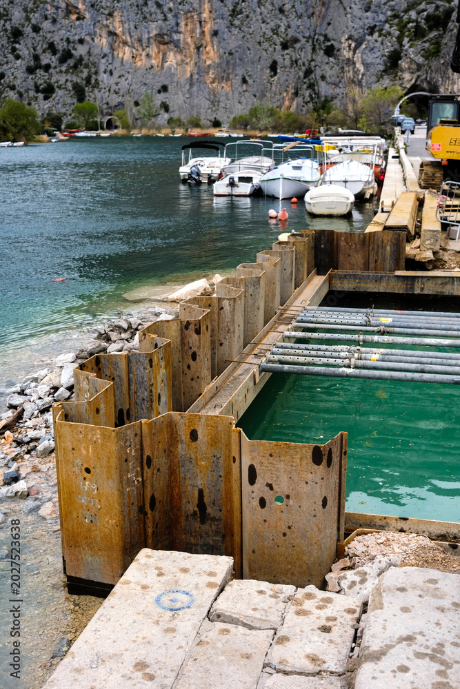 Obraz premium Sheet piling creates a dry work area for harbor development on a waterfront, with boats docked nearby and a dramatic rocky landscape beyond